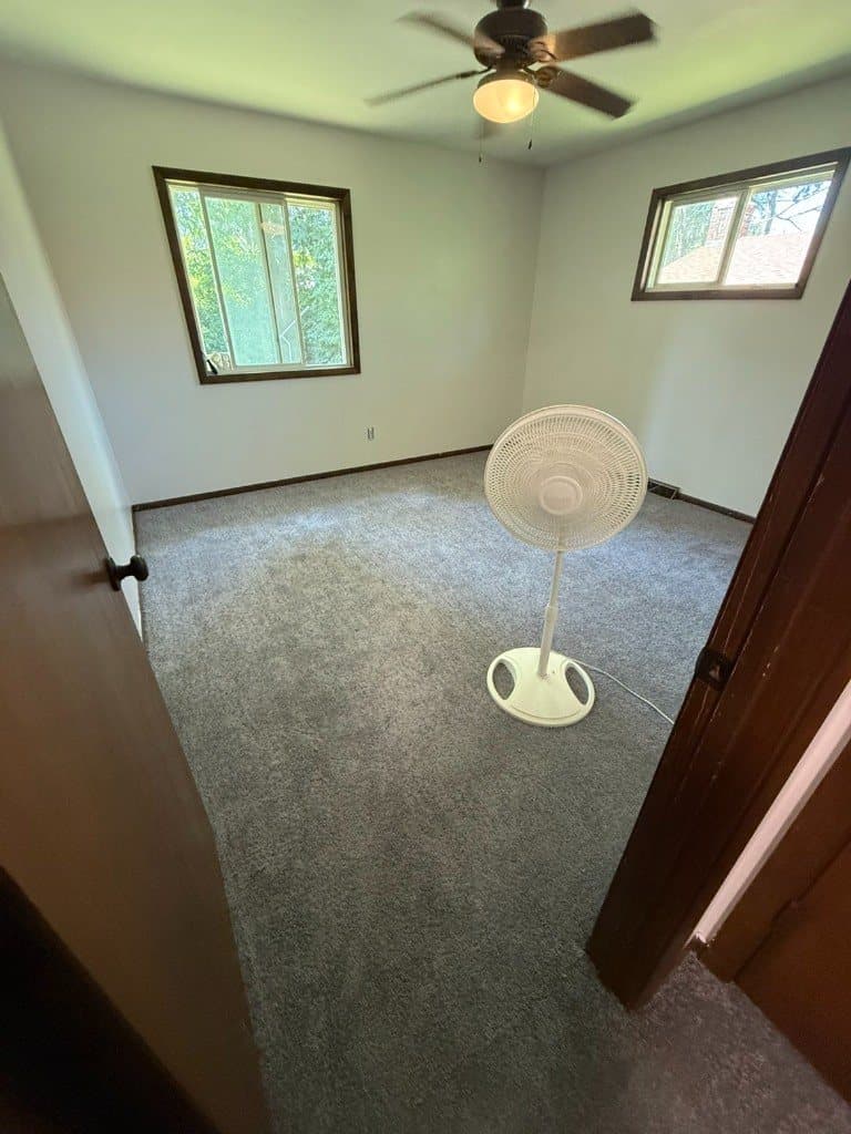 Gray carpet in an empty bedroom with two windows, a ceiling fan, and a white pedestal fan