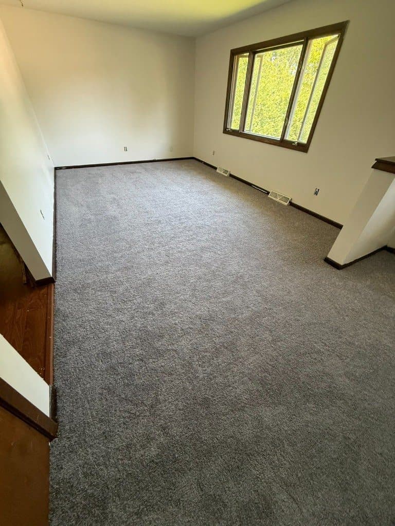 Gray wall-to-wall carpet in an empty living room with a wide window and wood trim